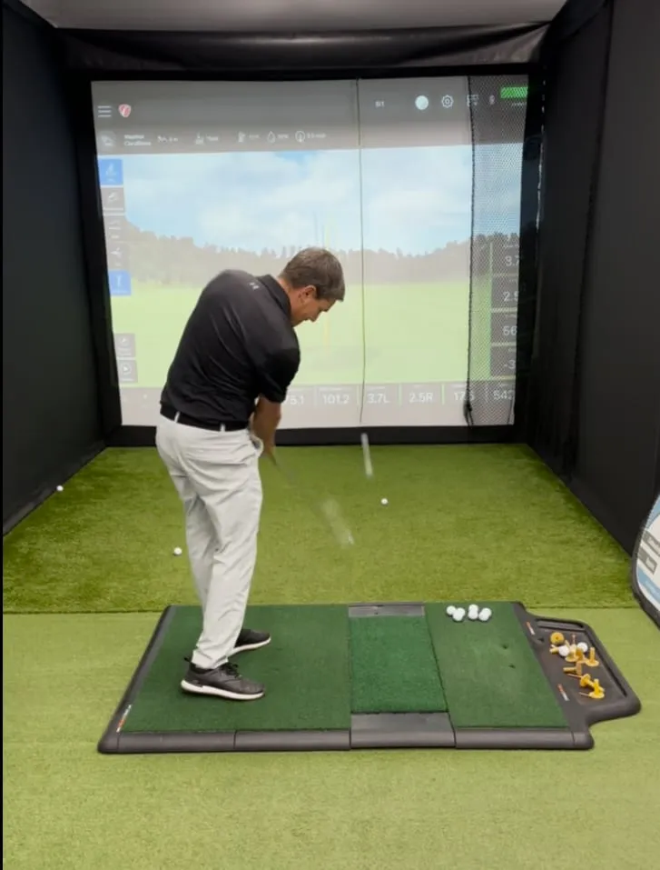 A club fitting session in a simulator bay, launch monitor numbers on the screen mid-shot
